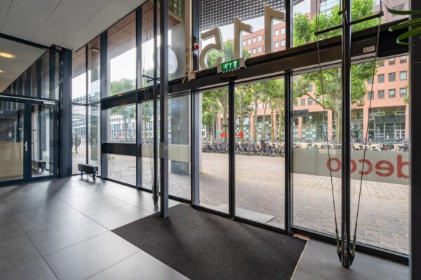 Glazen entreedeuren van een modern gebouw aan het Johanna Westerdijkplein met uitzicht op een plein vol geparkeerde fietsen.